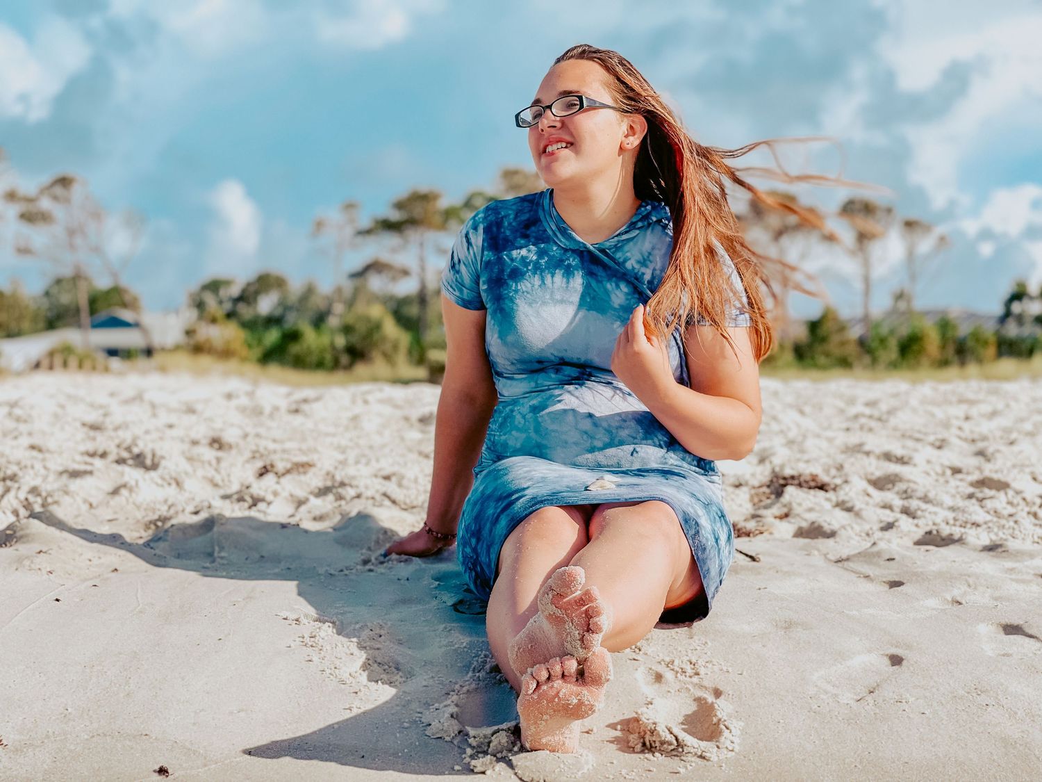 A joyful individual in tie-dye dress sits on a sandy beach with windswept hair and bare feet on a sunny day.