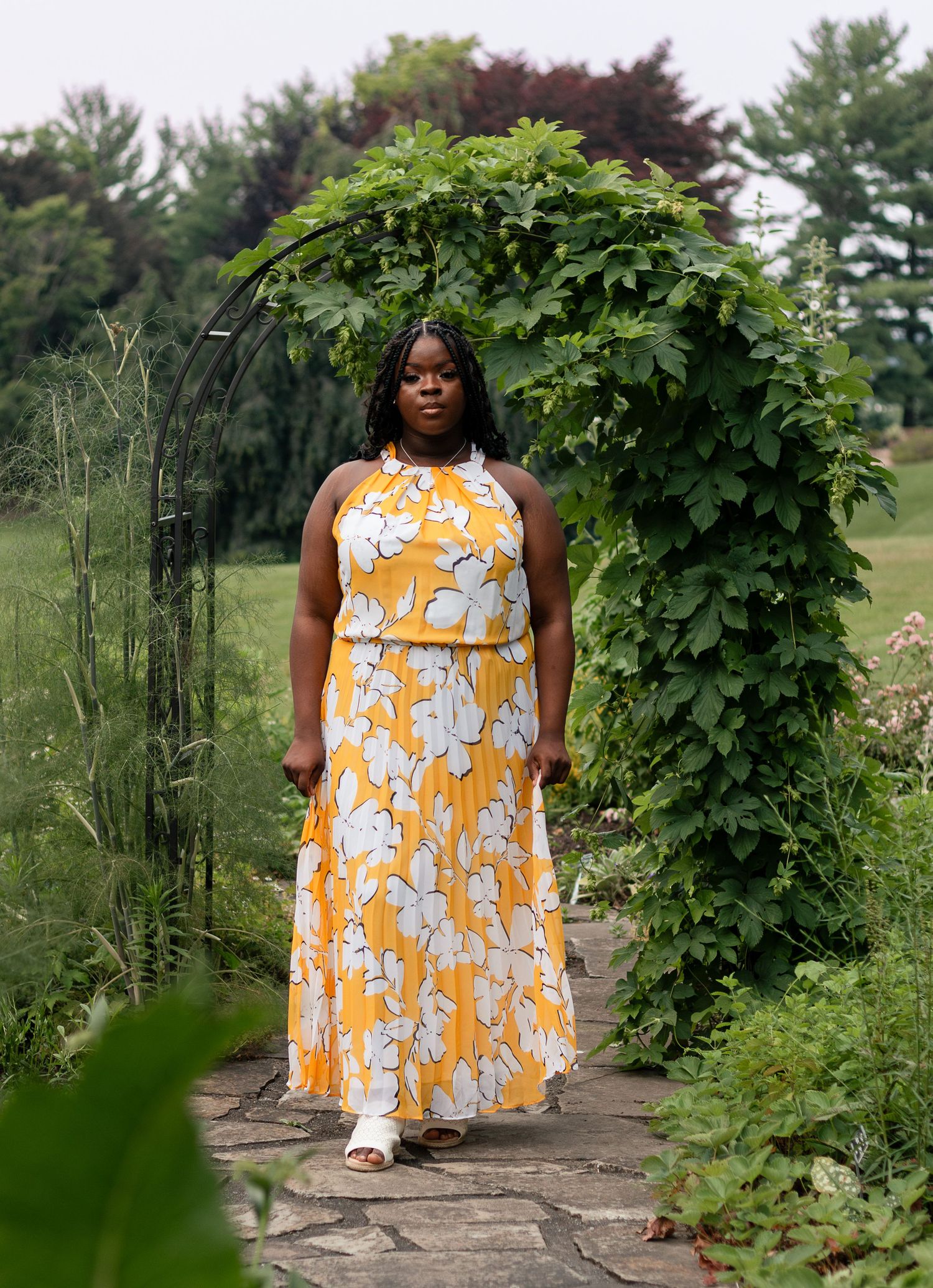 A person in a vibrant yellow floral maxi dress stands in a garden path beneath a lush green vine archway.