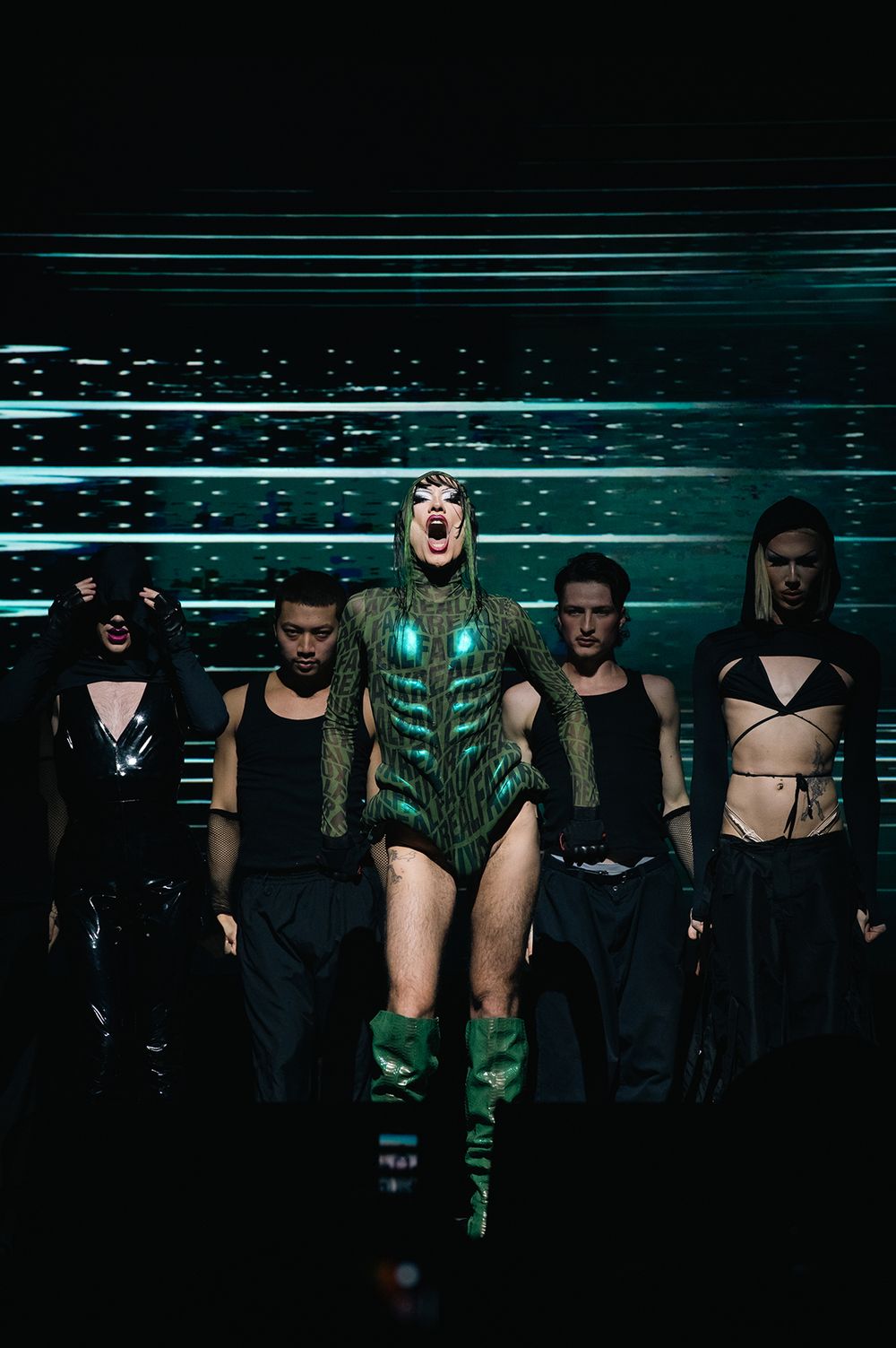 Theatrical drag performance with dancers in dark costumes against teal lighting effects.