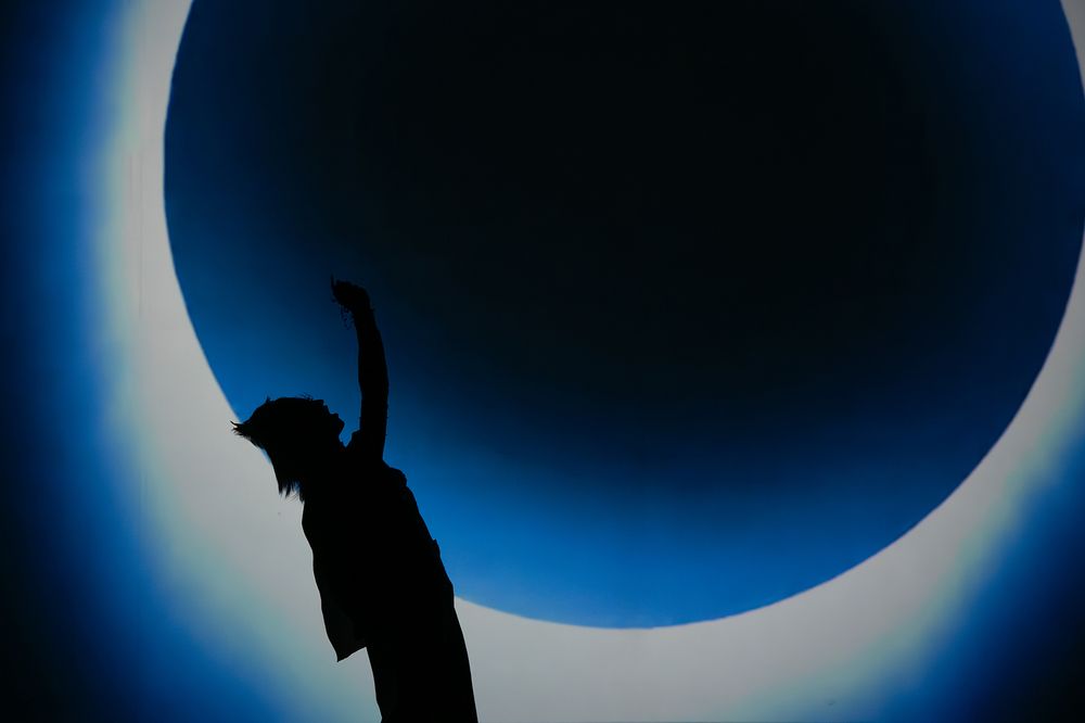 Silhouetted figure stands against dramatic blue circular light creating an ethereal eclipse-like effect.