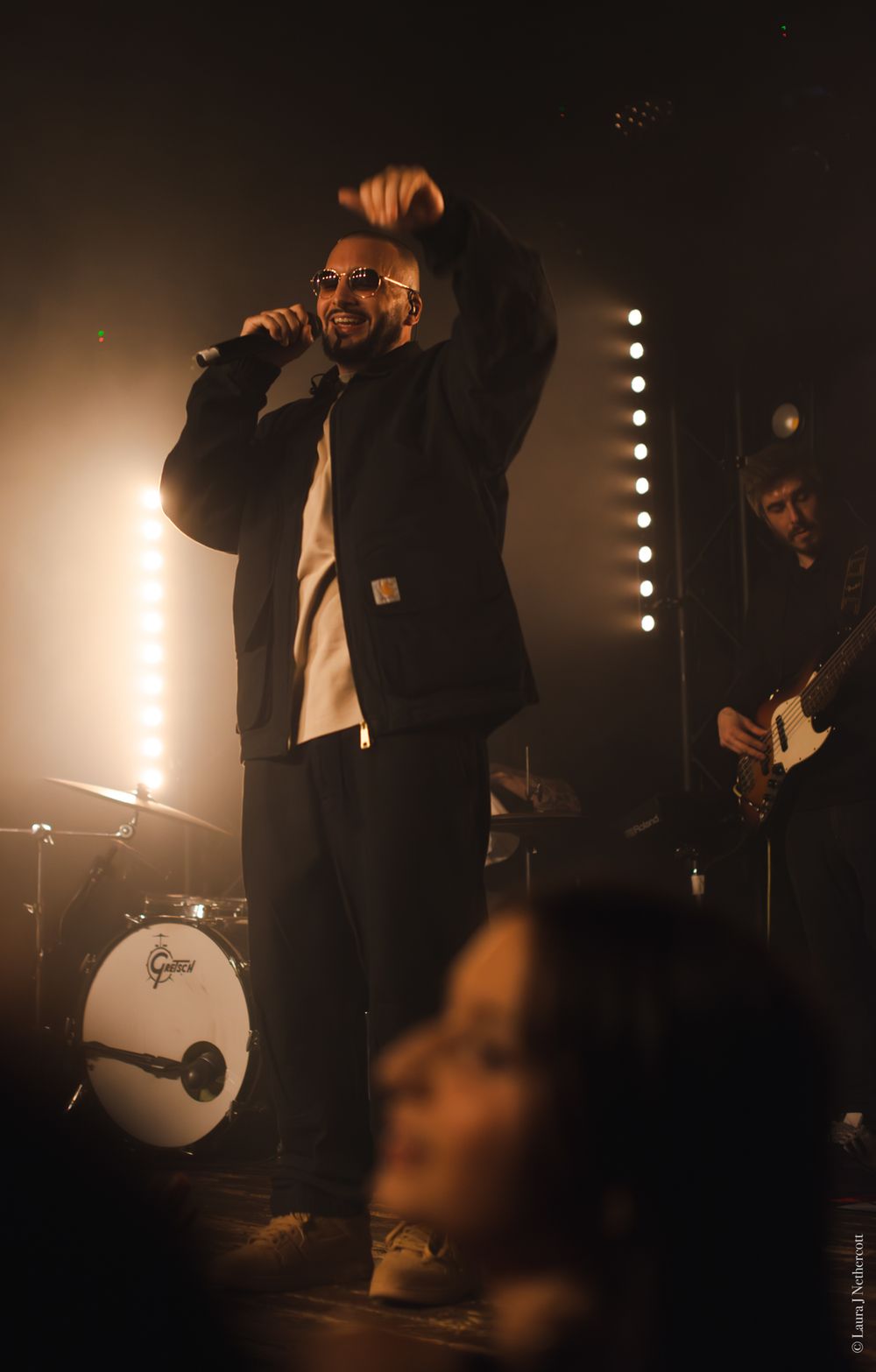 Concert performer in black jacket singing on stage with warm lighting and drum kit visible.