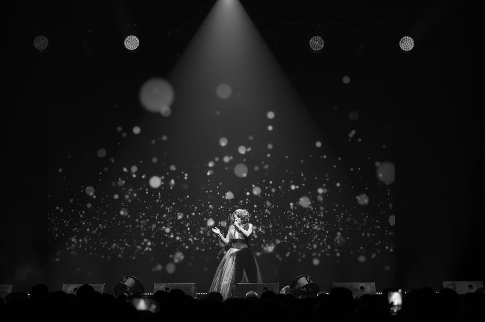 Dramatic black and white concert photo with performer surrounded by sparkles and spotlight effects.