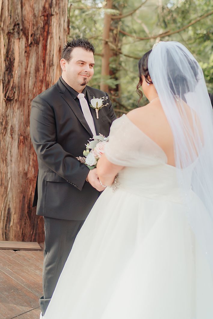 A bride and groom share a romantic first look moment in a sun-dappled forest setting.
