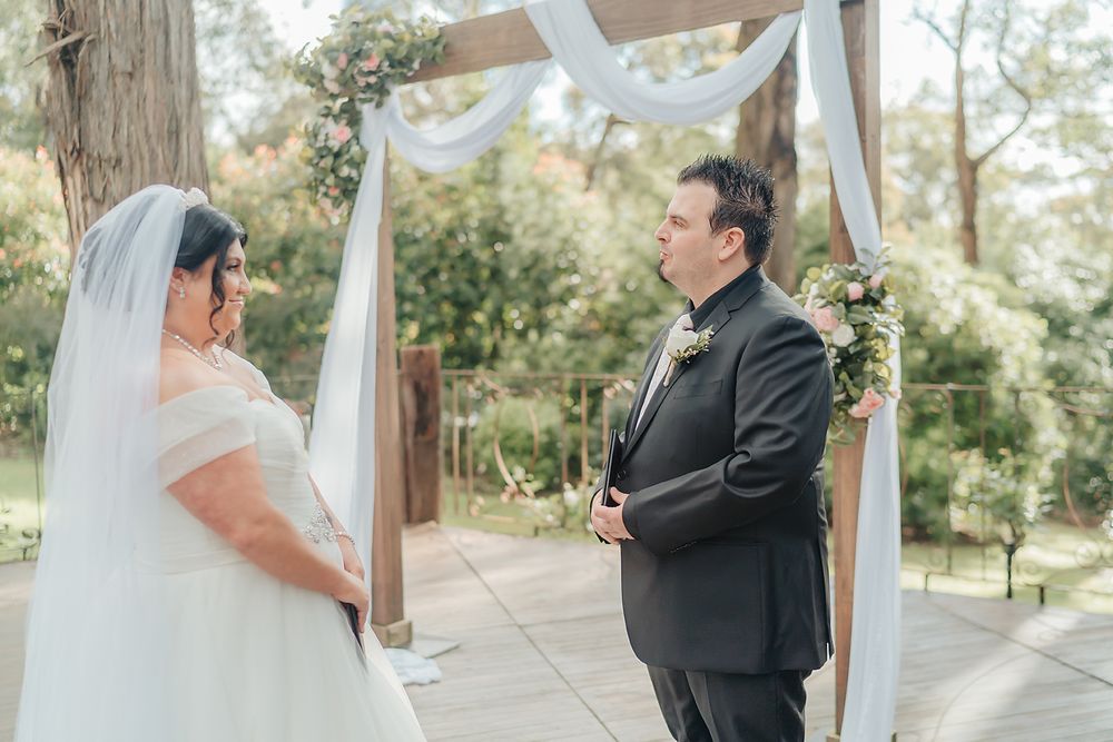 A touching outdoor wedding ceremony under a white draped arch surrounded by trees and natural scenery.