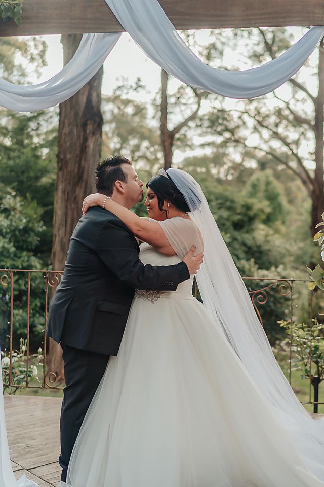A romantic outdoor wedding scene with decorative white drapes and a couple embracing under a beautiful wooded canopy.