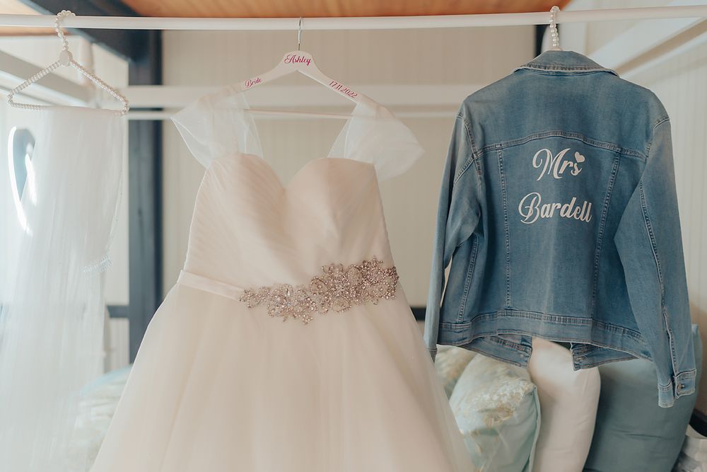 A white wedding dress with crystal embellishments hangs next to a custom denim jacket with 'Mrs' embroidery.