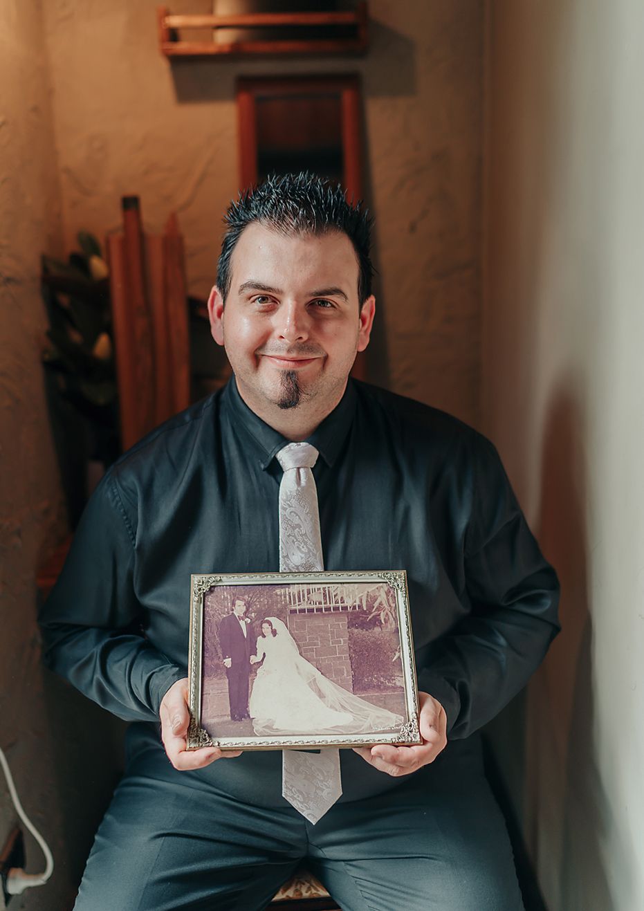 A person in a dark shirt and tie holds up a vintage black and white wedding photograph in a warm-lit indoor setting.