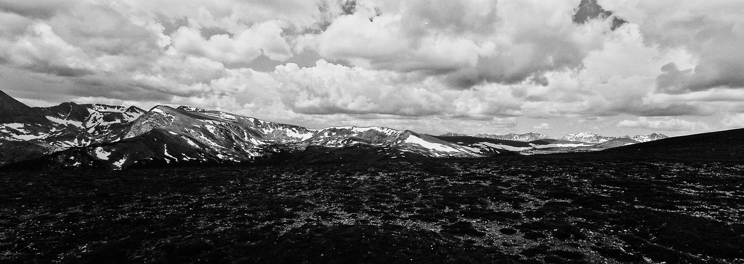 Black and white landscape of snow-capped Rocky Mountains under dramatic cloudy skies.