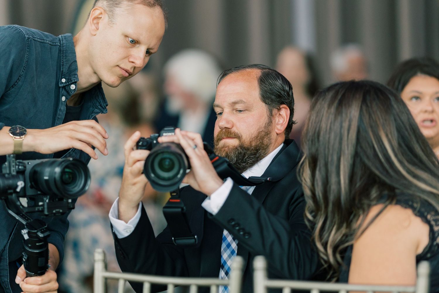 Professional photographer in formal attire examines camera settings while seated at an event with cinematographer nearby.