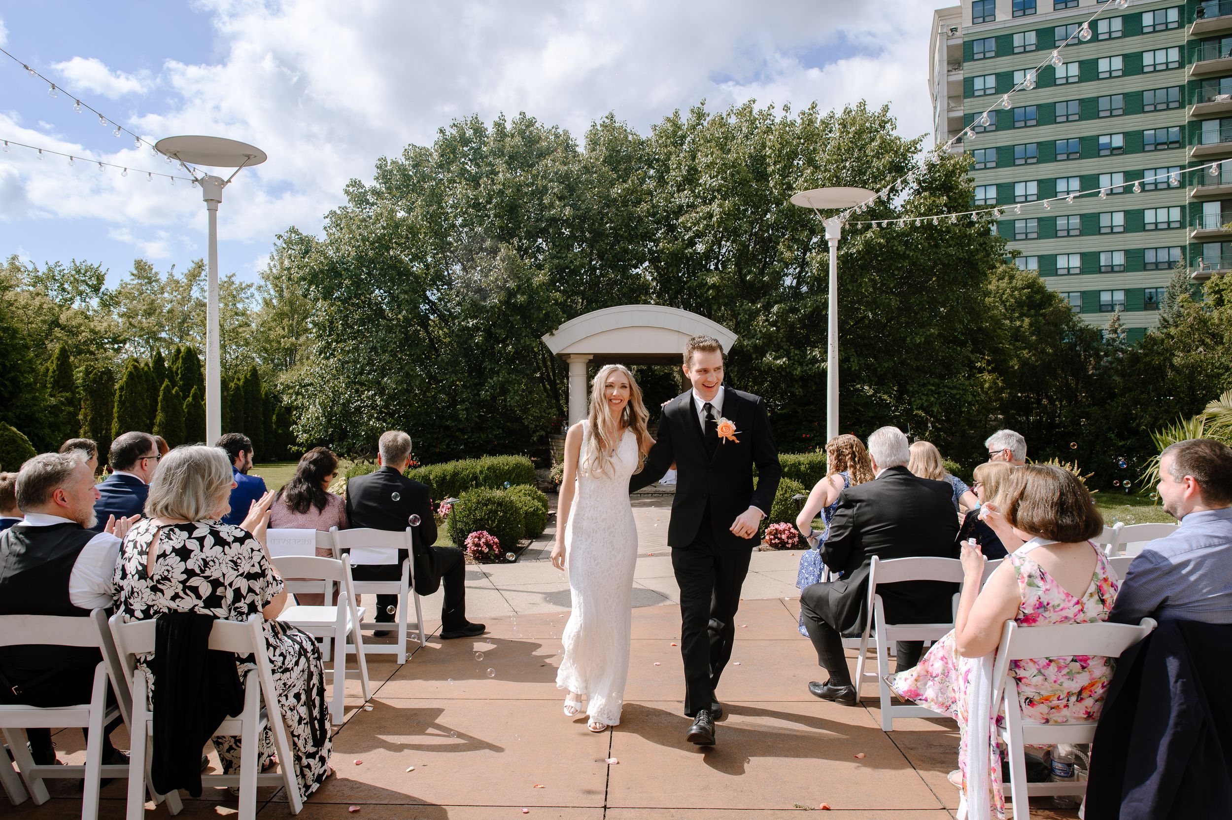 Wedding guests gather on an outdoor terrace at a seaside venue under a cloudy sky and string lights.