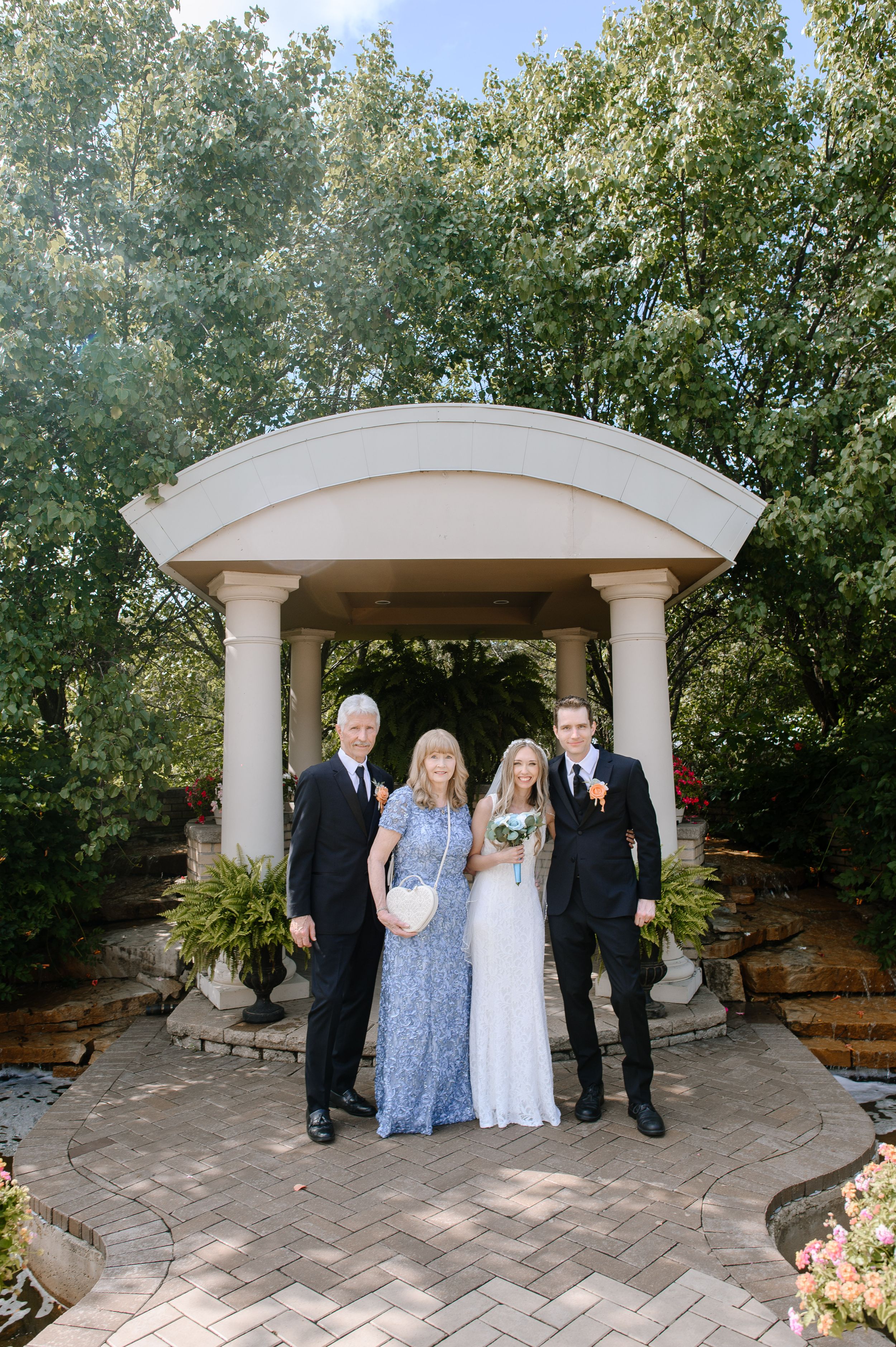 A wedding party poses under an elegant outdoor gazebo surrounded by lush greenery and stone pavers.