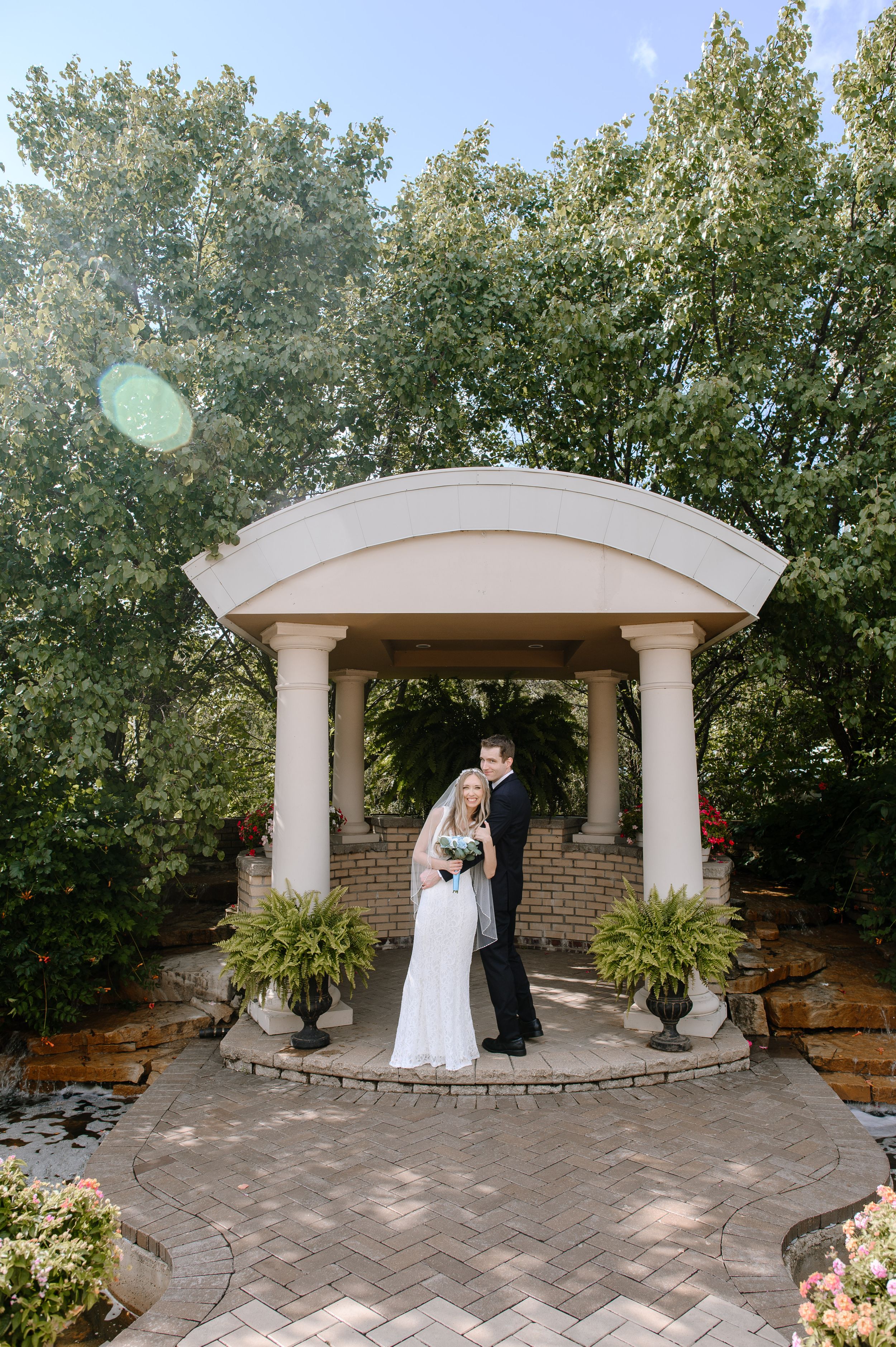 A newlywed couple stands together under an elegant white garden gazebo surrounded by lush greenery and stone pavers.