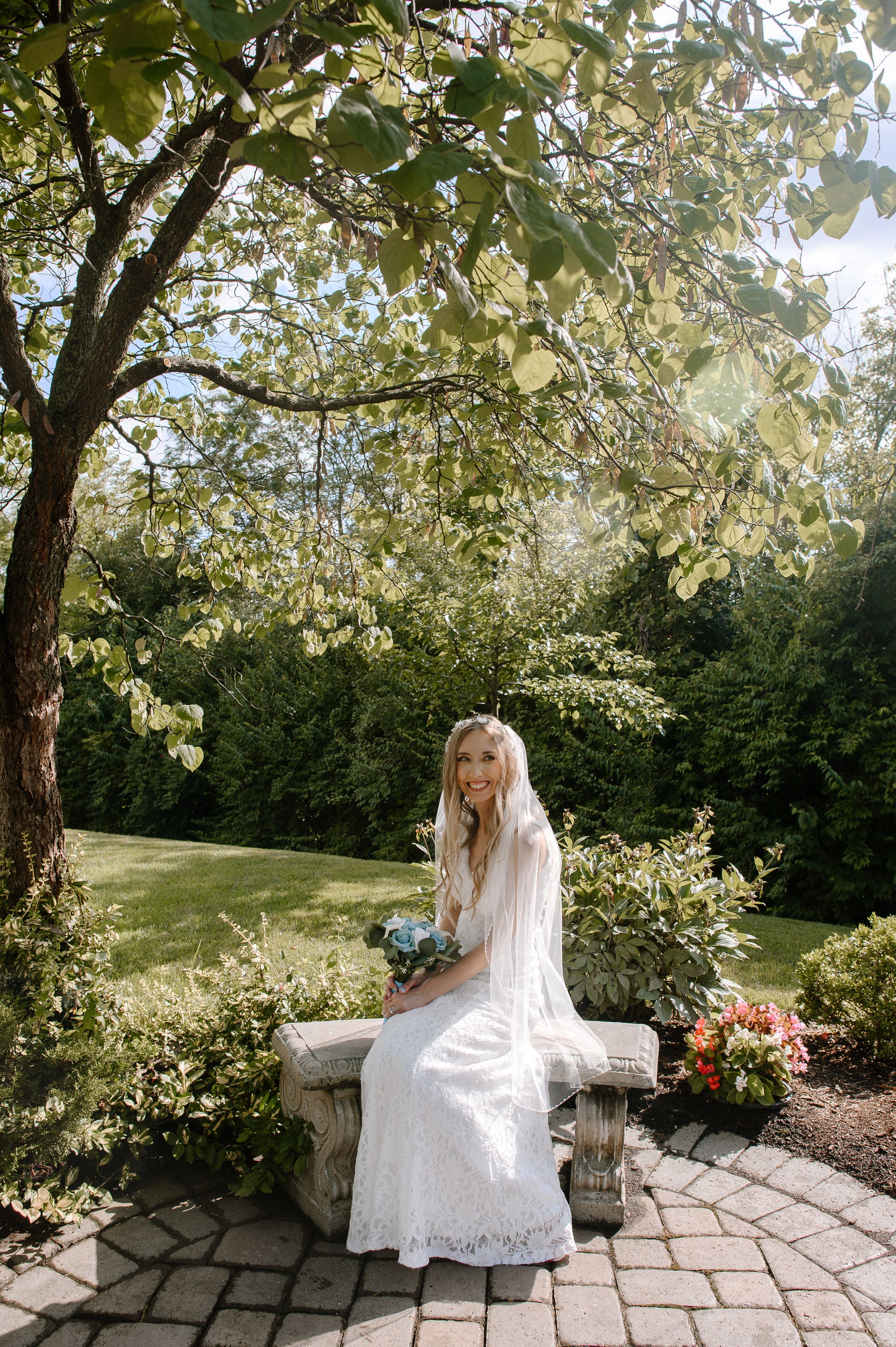 A bride in a white lace dress sits on a stone bench in a garden setting with flowering trees and sunlight.