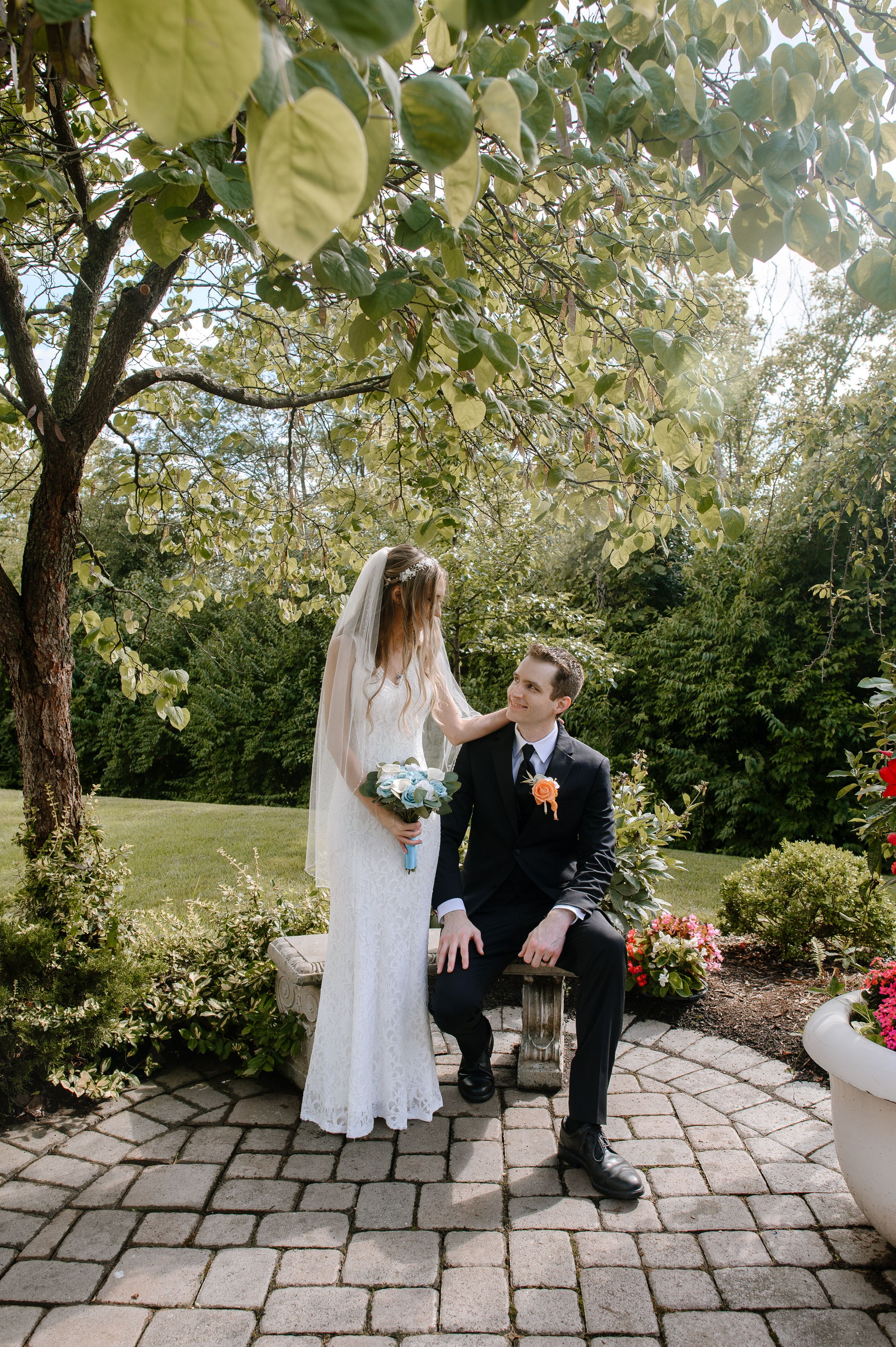Wedding couple sits together on stone bench in lush garden surrounded by blooming trees during outdoor ceremony.