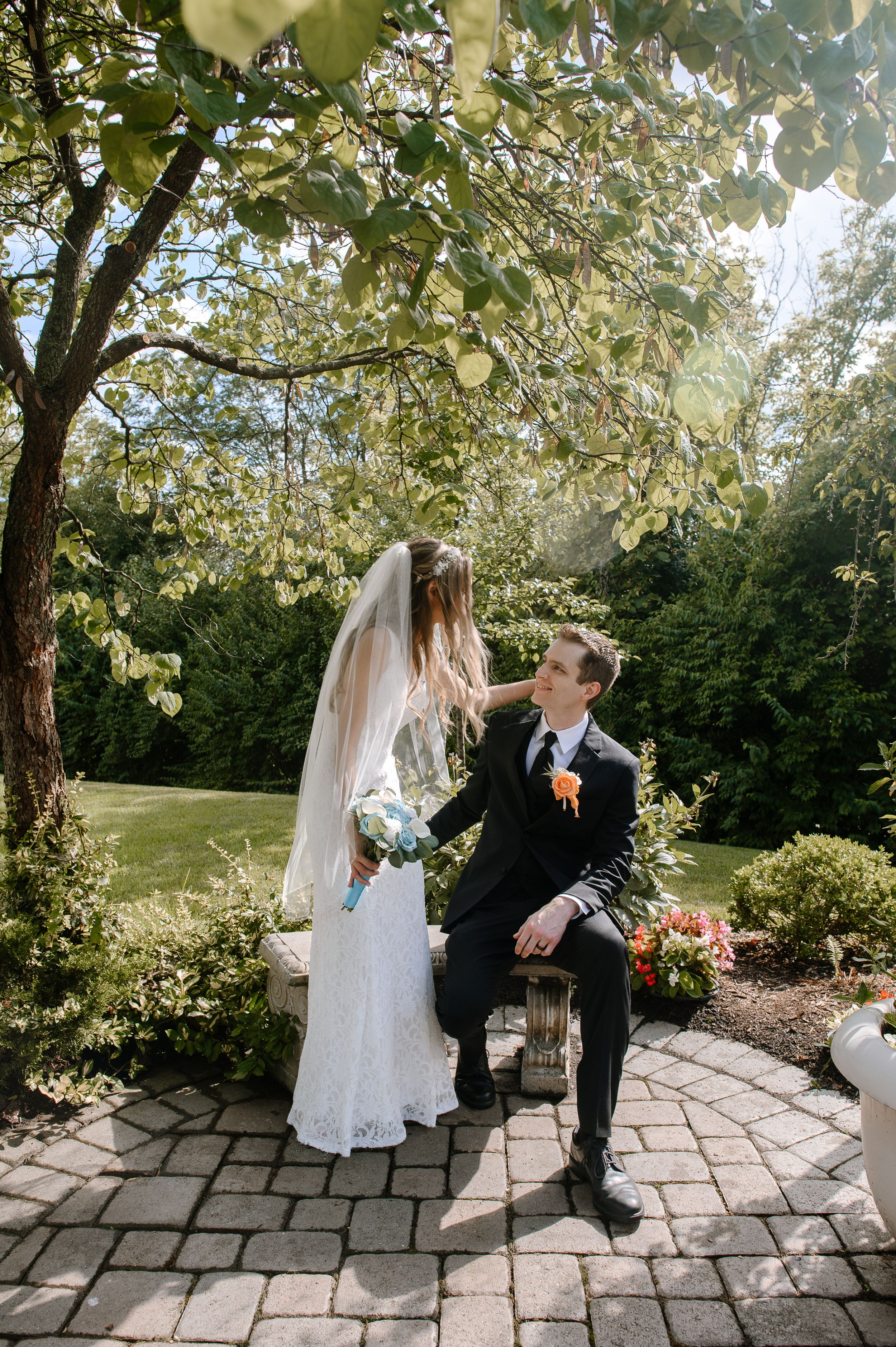 A couple share a romantic moment on their wedding day in a sunlit garden with blooming trees overhead on a stone patio.