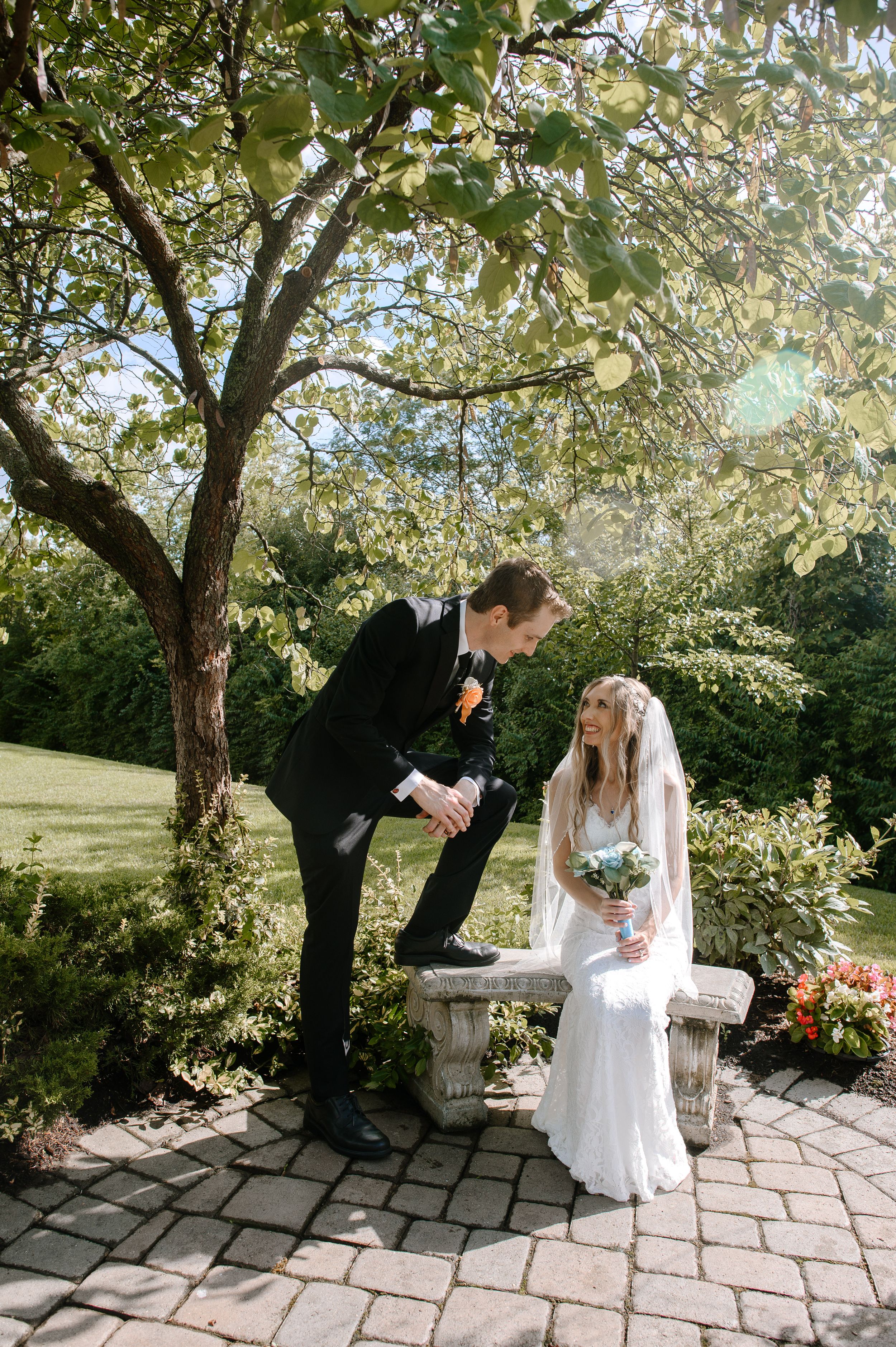 Newlyweds share a tender moment by a stone bench in a sunlit garden setting.