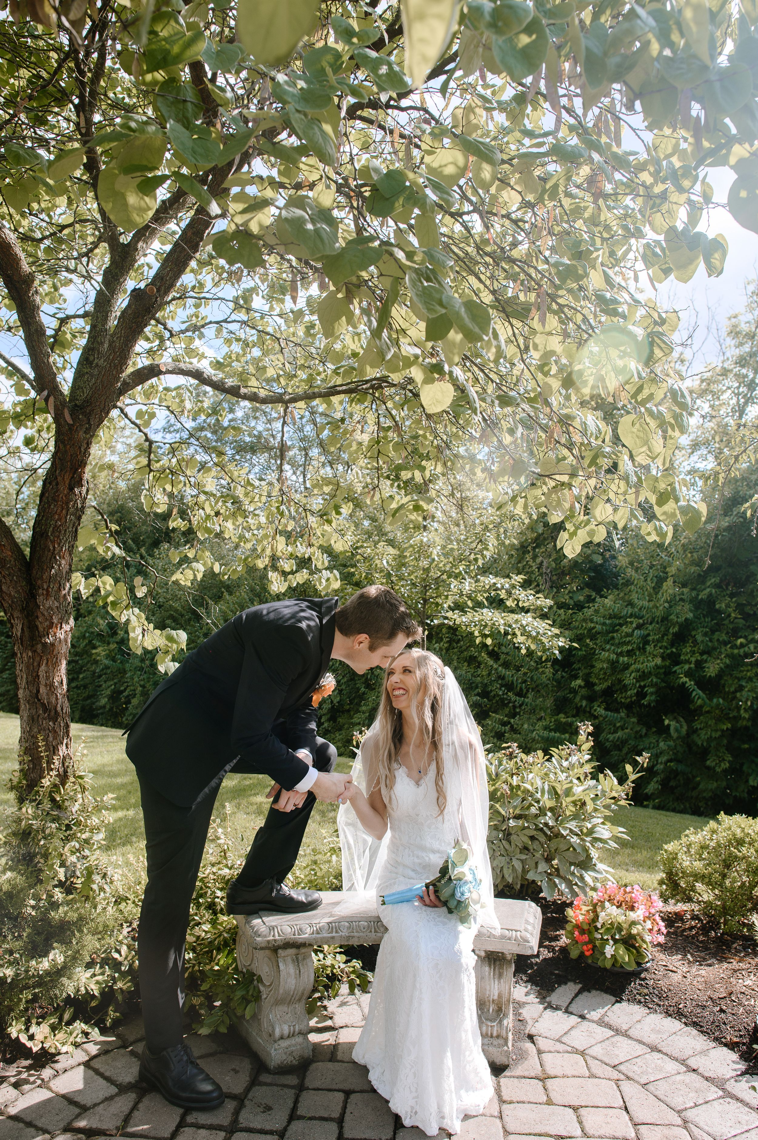 A romantic wedding photoshoot near a stone bench under blooming trees in a garden setting.