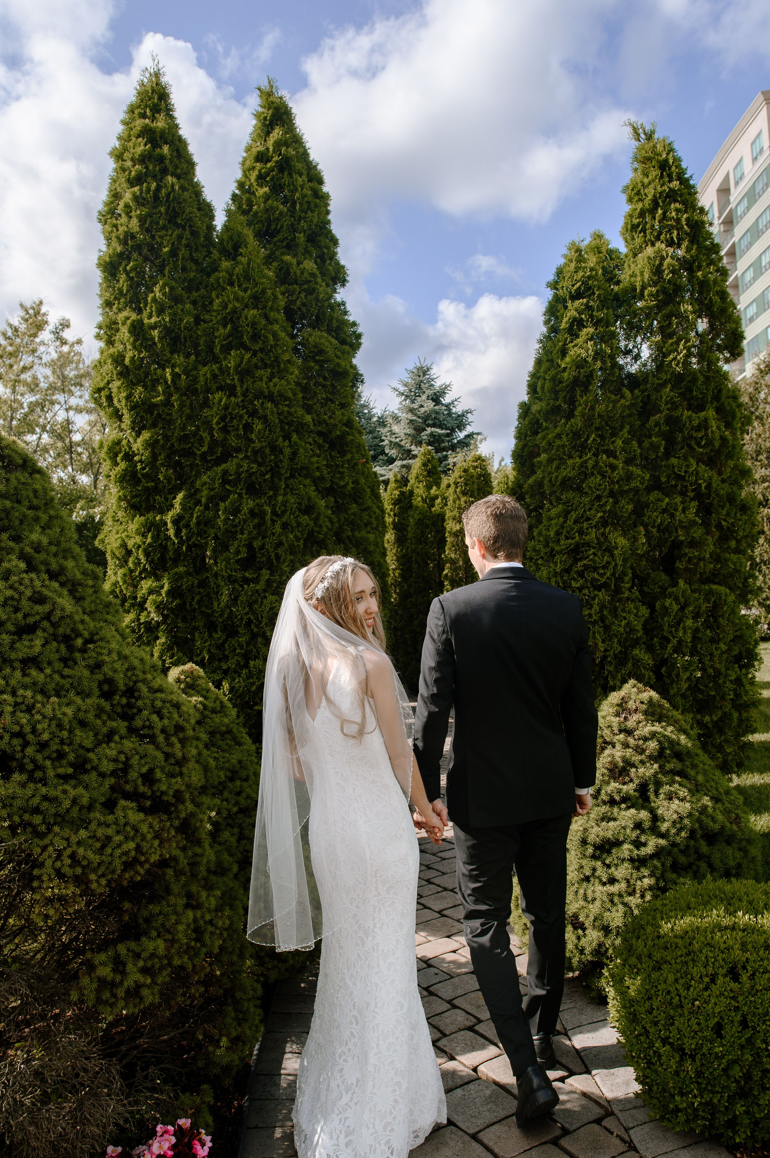 A couple walks hand in hand on a stone pathway surrounded by tall evergreen trees and shrubs in a garden setting.