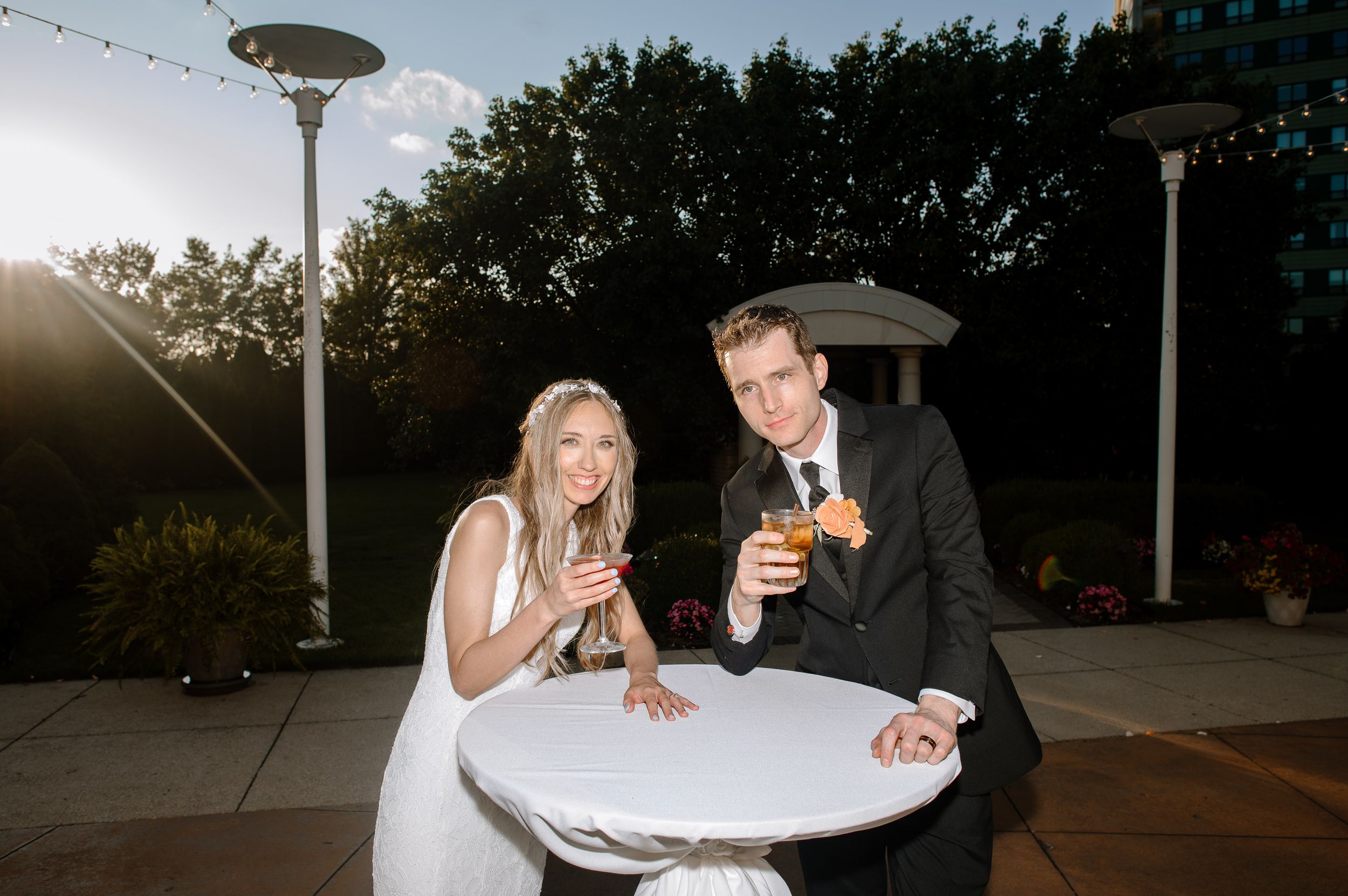 A couple at an outdoor bistro table sharing drinks and laughing at sunset with string lights overhead.