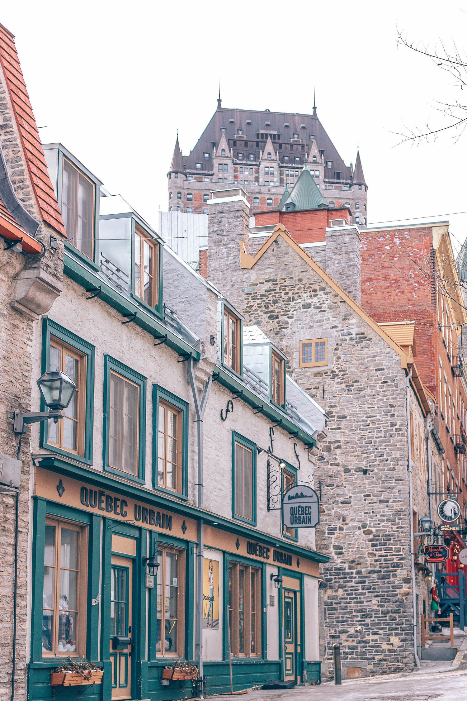 Historic stone buildings with teal trim line a street in Old Quebec City with Chateau Frontenac tower visible in background.
