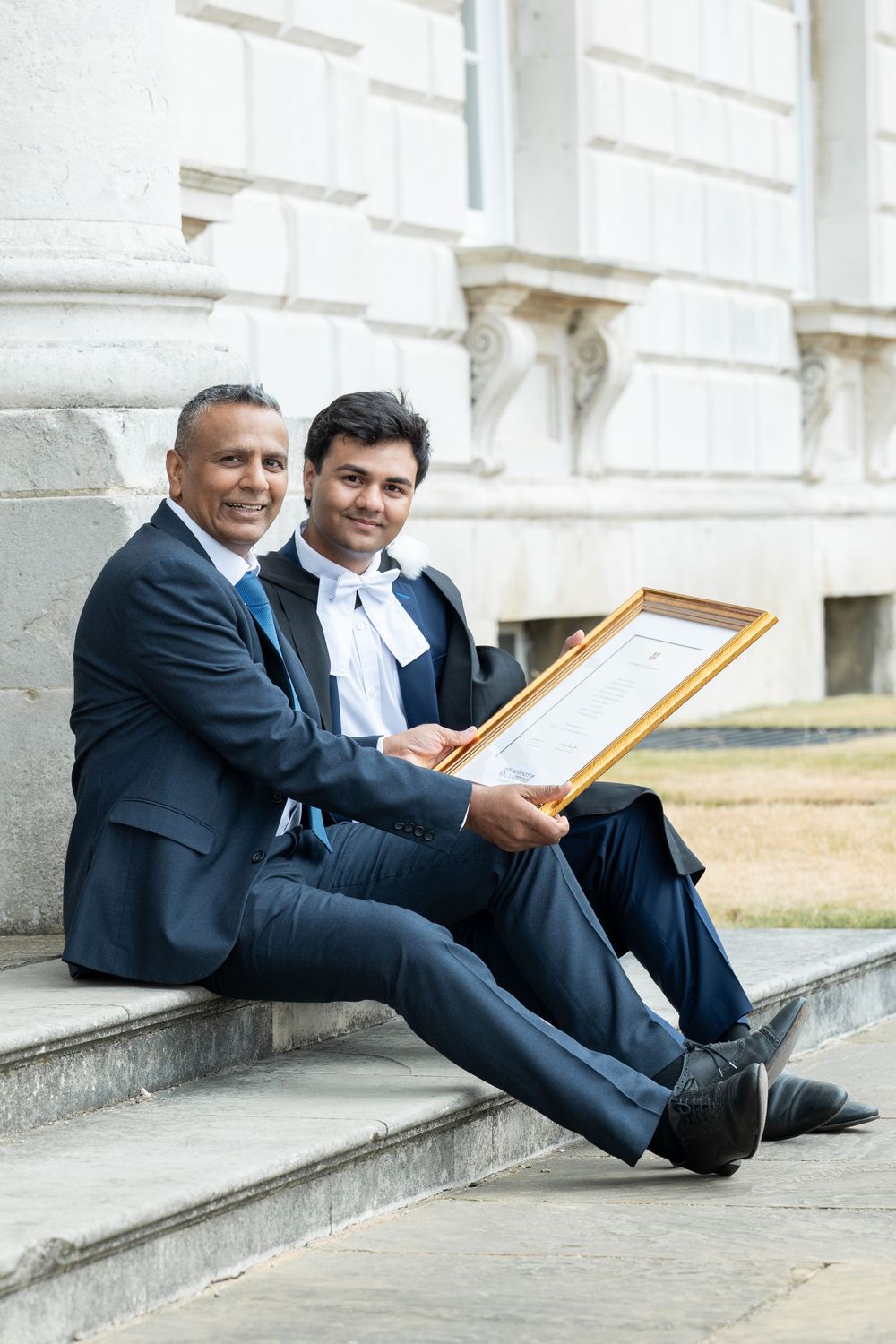 Two people in business suits sit casually on outdoor stone steps while reviewing documents.
