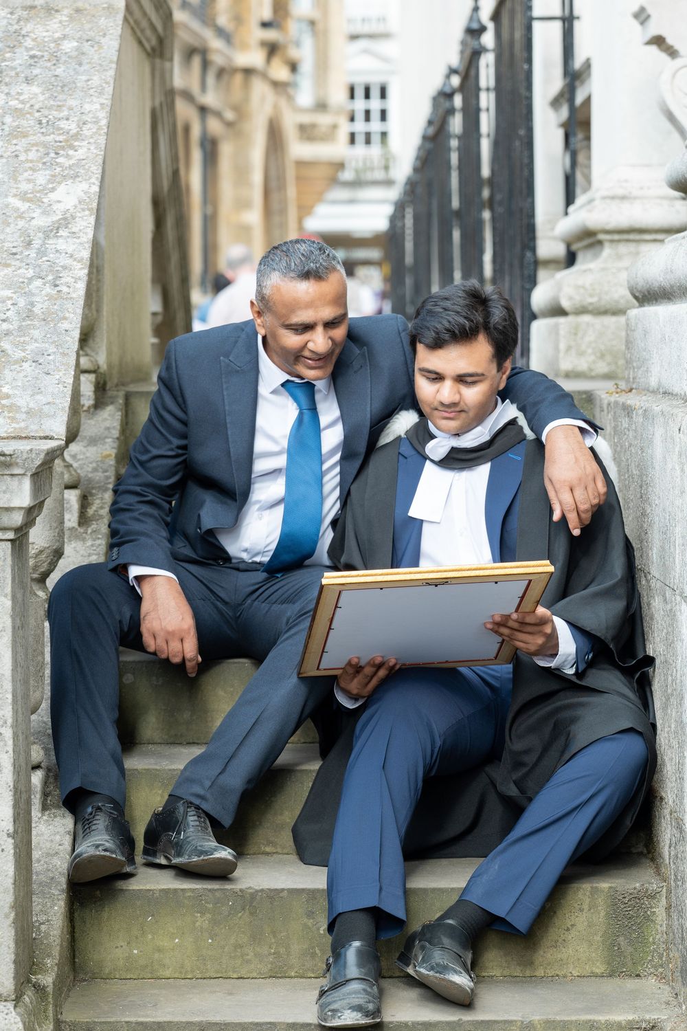 Two professionals in business suits sit together on stone steps reviewing documents with a tablet or folder.