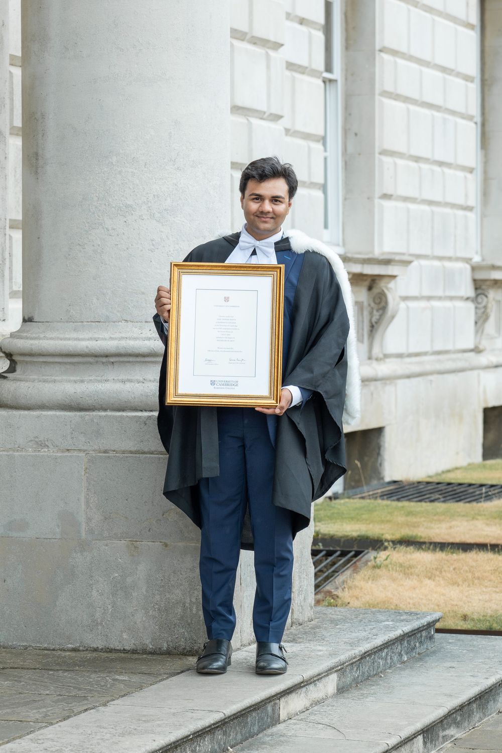 A graduate in academic regalia stands on stone steps holding up a framed diploma certificate outside a classical building.