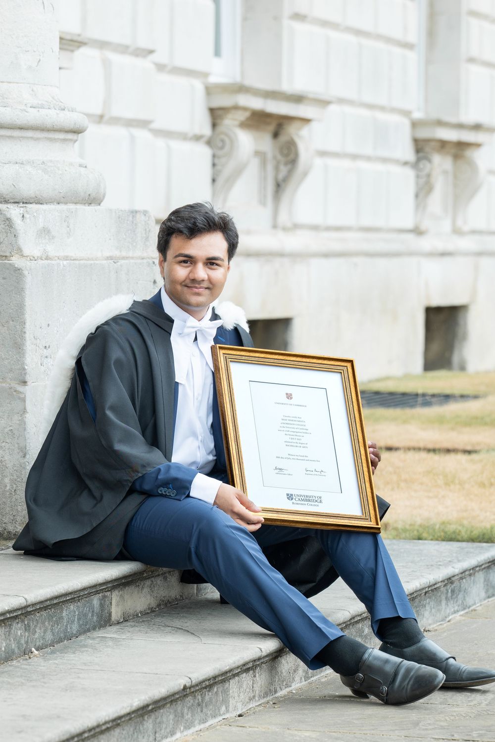 Graduate in academic dress sits on stone steps holding framed diploma certificate in gold frame with white building behind.