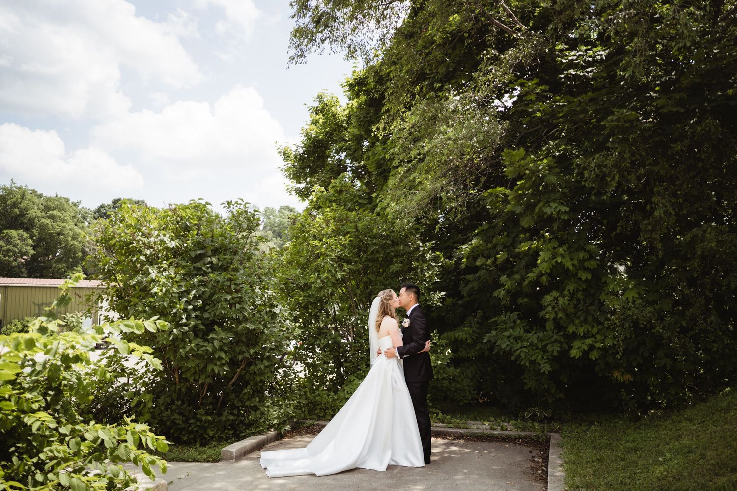 A couple in wedding attire embrace on a stone pathway surrounded by lush summer greenery and trees.