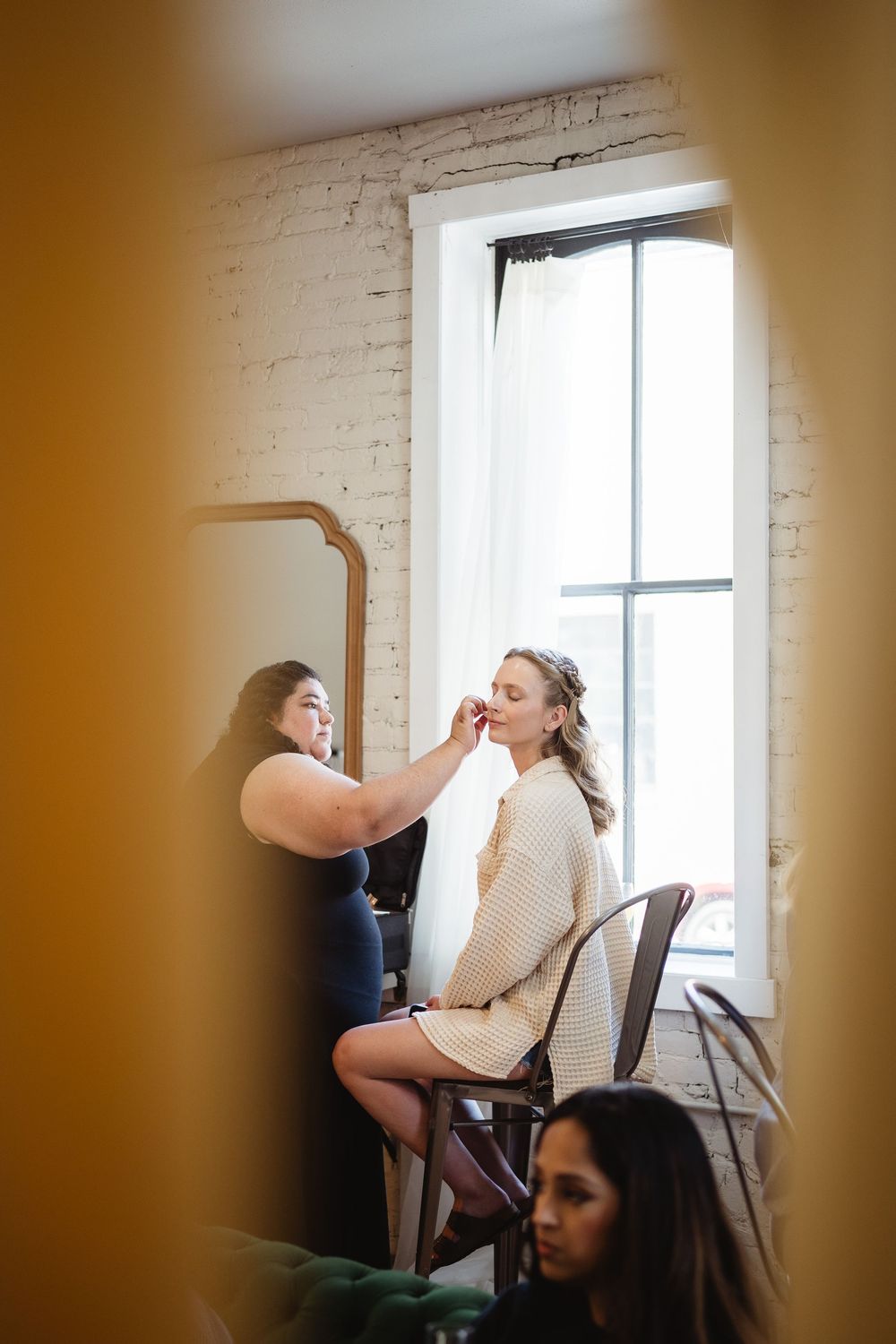 A makeup artist applies cosmetics to a person seated in a chair near a bright window in a brick-walled room.