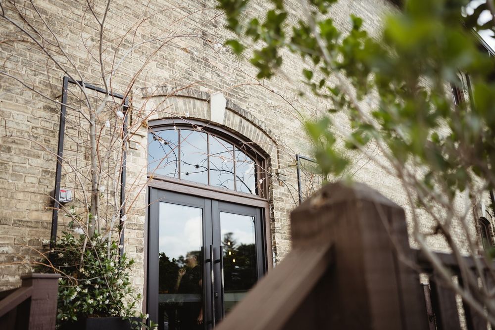 An arched entrance to a historic brick building with glass doors and decorative plants framing the doorway.