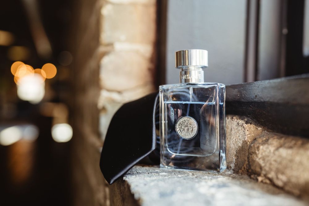 A glass perfume bottle with silver cap sits on a stone ledge with blurred bokeh lights in the background.