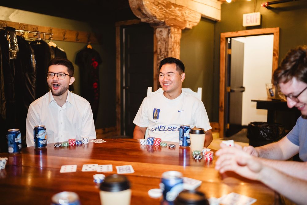 Three people sitting at a wooden table with cans of beverages in what appears to be a casual bar or meeting space.