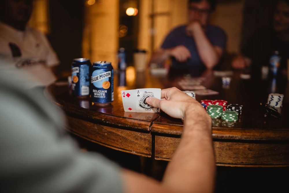 People play cards while enjoying drinks at a wooden bar counter in dim lighting.