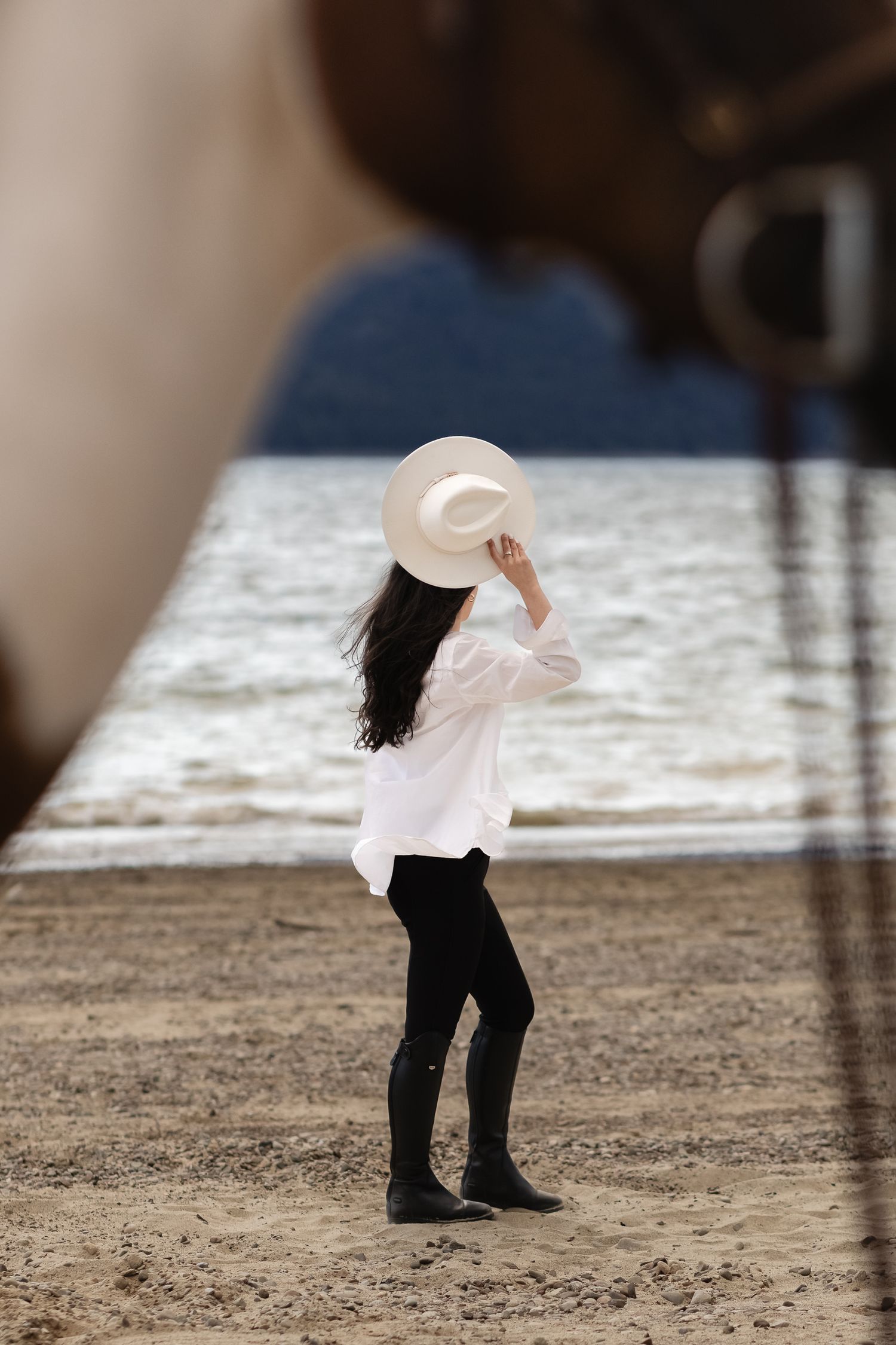A model holding a hat at Speelyi Beach is artistically framed by her horse in the foreground.