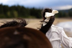 A woman with a white bow and her bay horse walking along Speelyi Beach with their hair blowing in the wind.