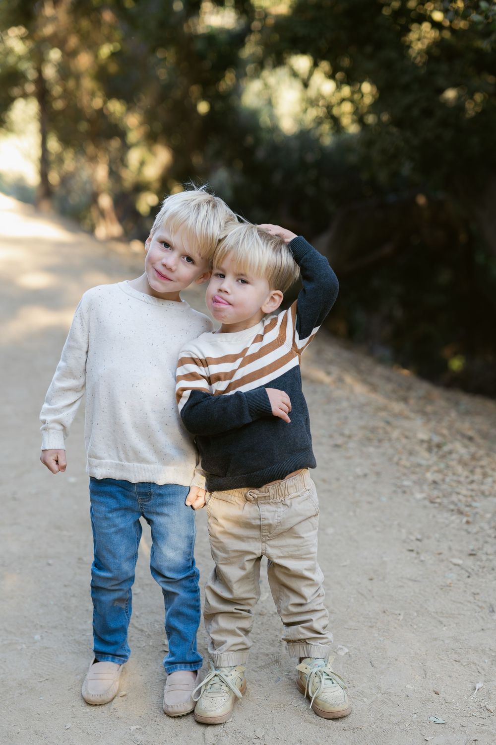 Two young children stand together on a dirt path wearing casual sweaters and pants on a sunny day.