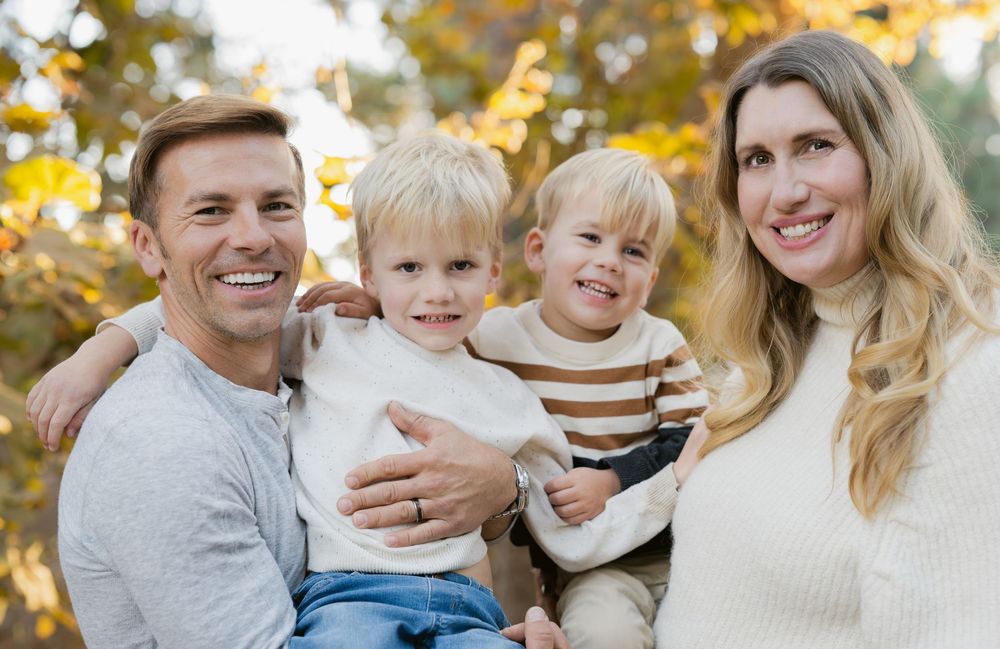 A family in coordinating grey and white outfits poses outdoors during autumn with golden leaves in background.
