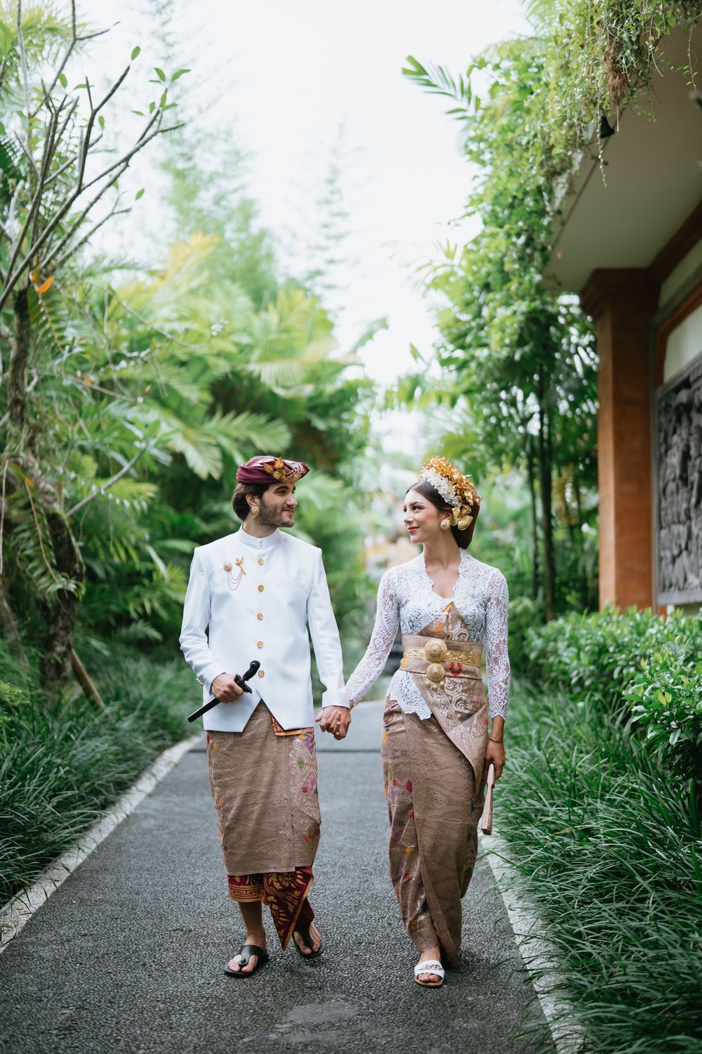 Two people in traditional dress walk along a garden path surrounded by tropical plants.