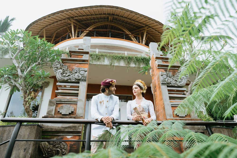 A couple poses on a wooden deck of a Balinese-style building surrounded by tropical foliage.