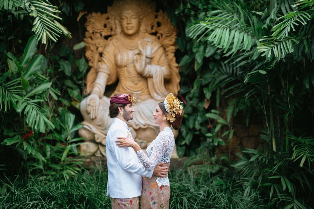 Two people in traditional Indonesian dress pose against a lush tropical garden backdrop.