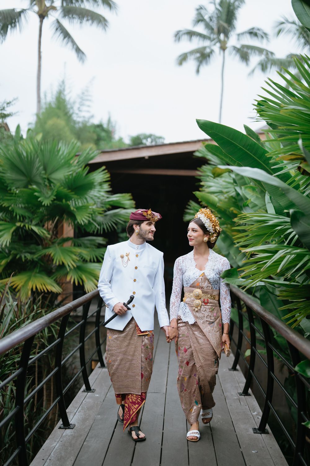 A couple in traditional Javanese wedding attire walks along a wooden pathway surrounded by tropical palm trees.