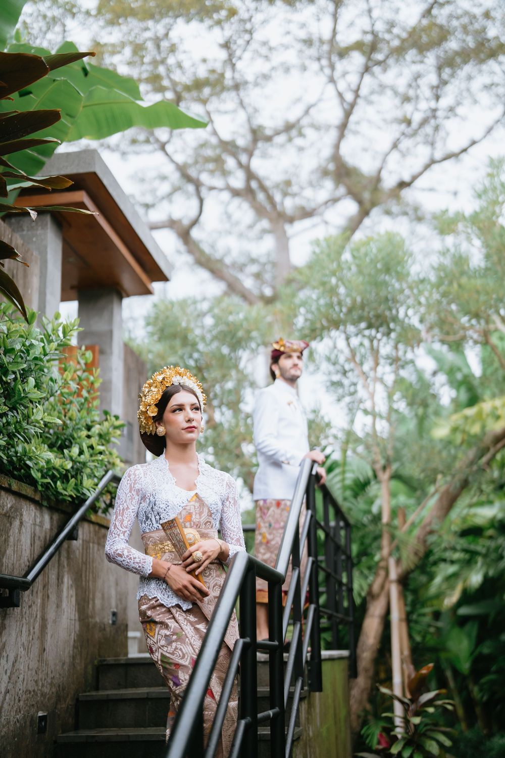 A person in traditional Indonesian dress poses on wooden stairs surrounded by greenery.