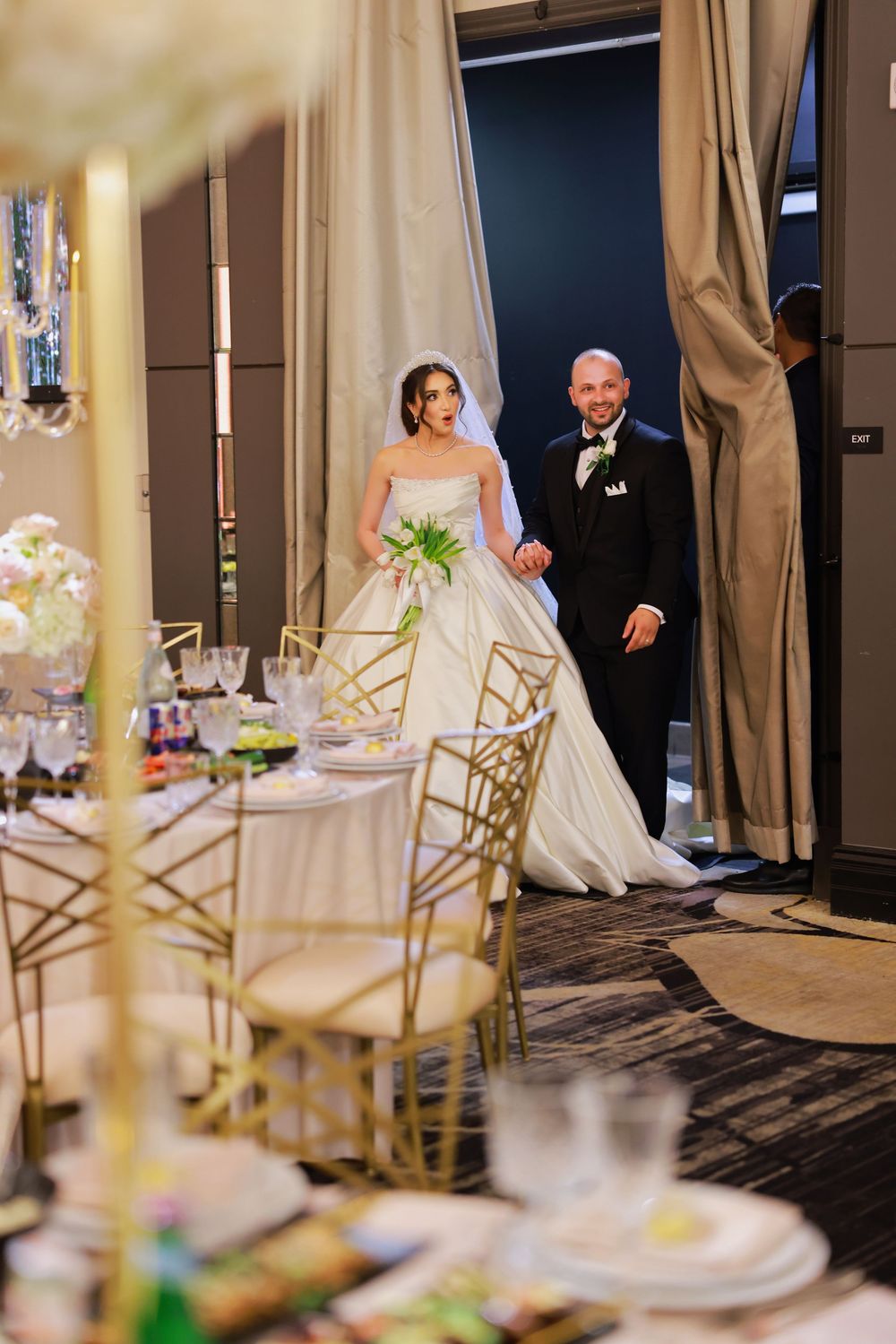 Wedding reception venue room with elegant table settings, gold chairs, and beige curtains.