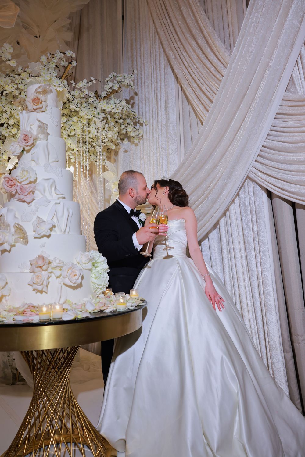 A romantic wedding scene with a stunning white draped backdrop and ornate floral cake display on a golden pedestal table.