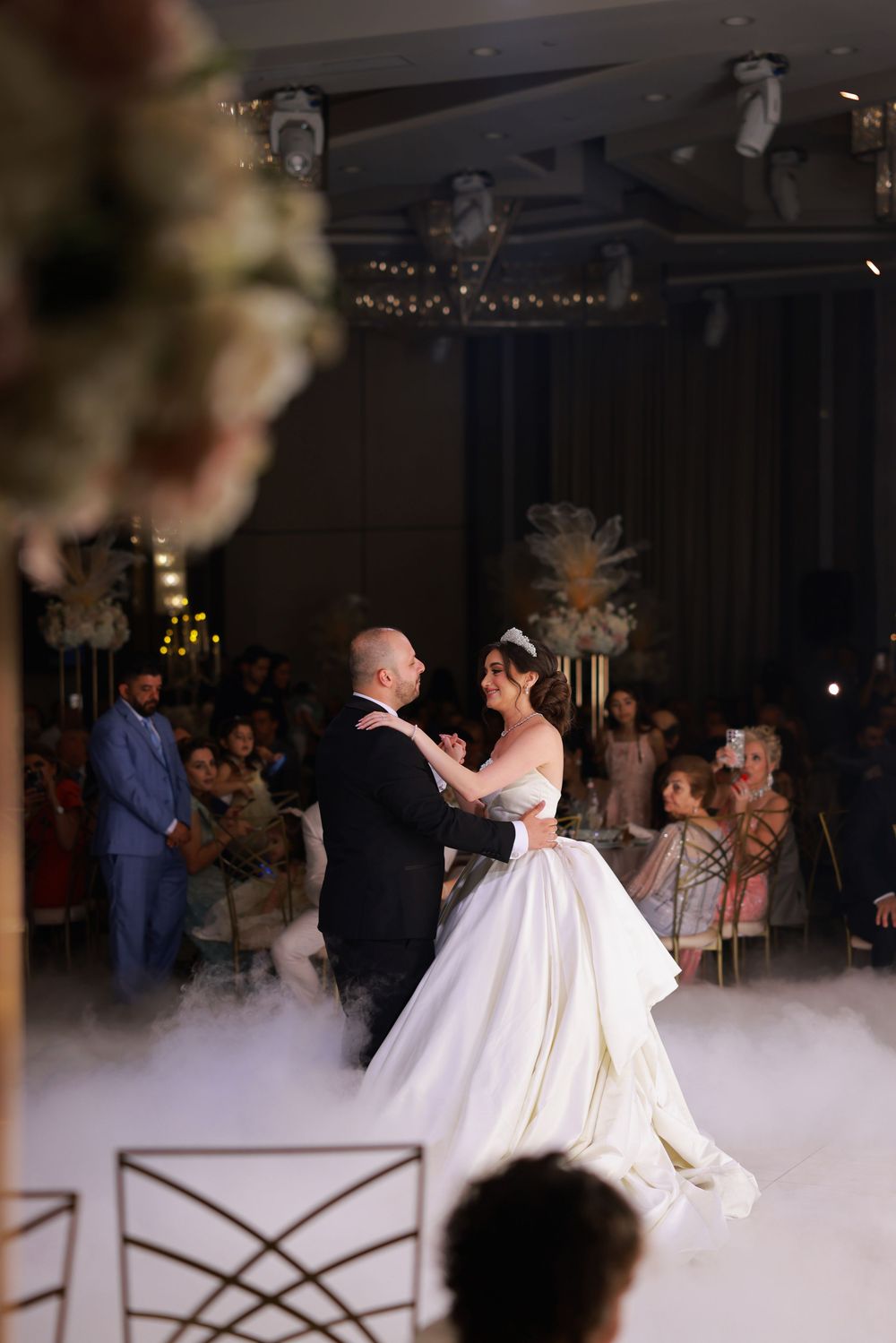 Couple shares their first dance on a misty dance floor with guests watching during an elegant wedding reception.