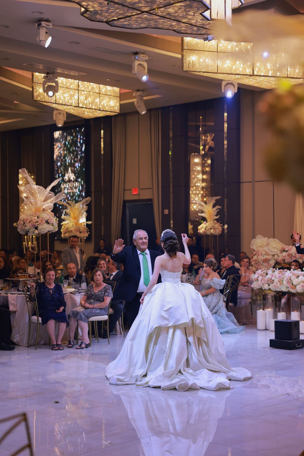 A couple dances on the white dance floor at an elegant wedding reception with crystal chandeliers overhead.