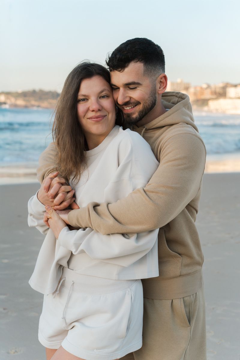 Romantic photoshoot with a couple hugging while standing on a sandy Bondi beach in Sydney at sunrise wearing neutral coloured clothing.