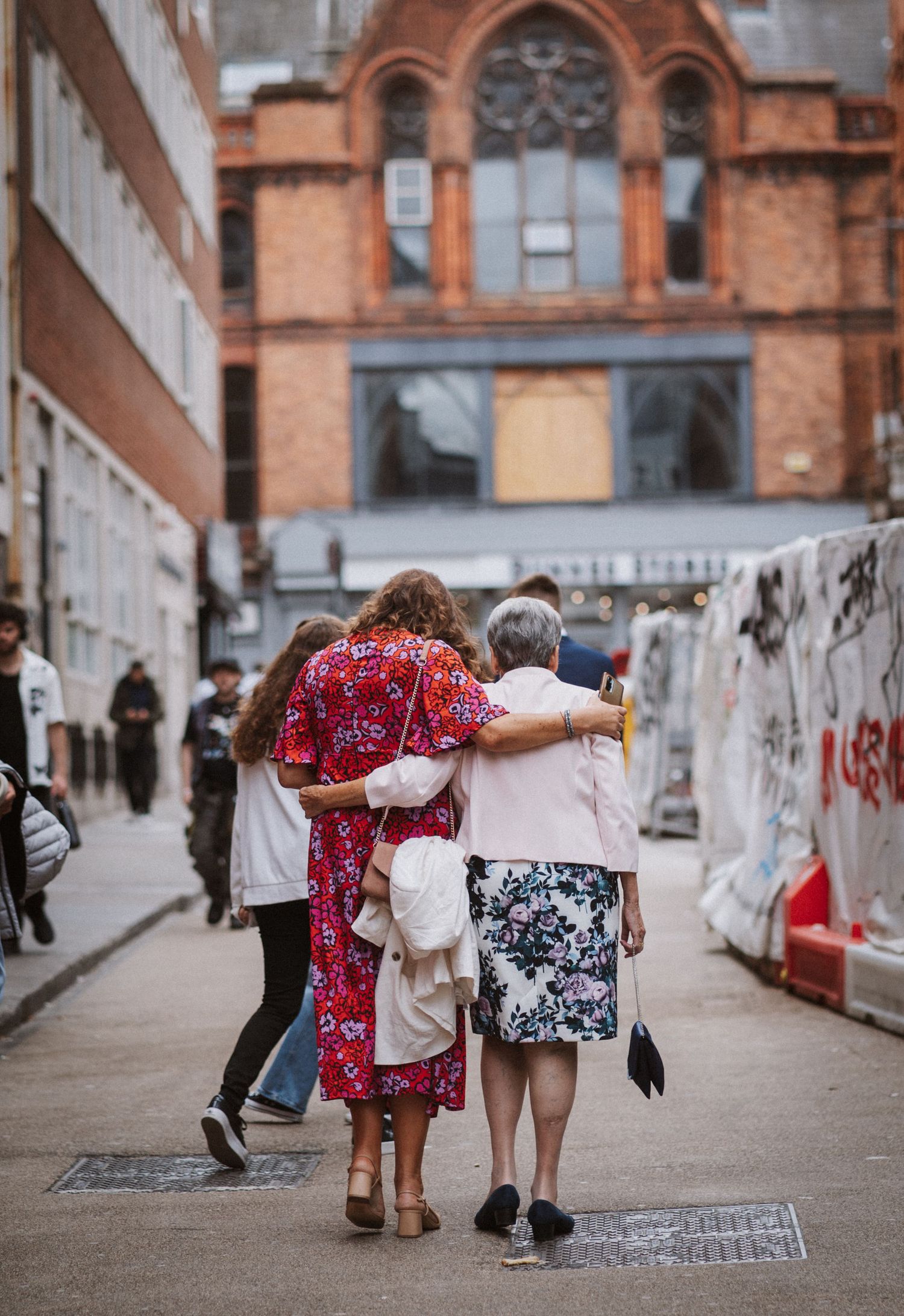 Two elderly women walk arm in arm down an urban street past graffiti walls and brick buildings.