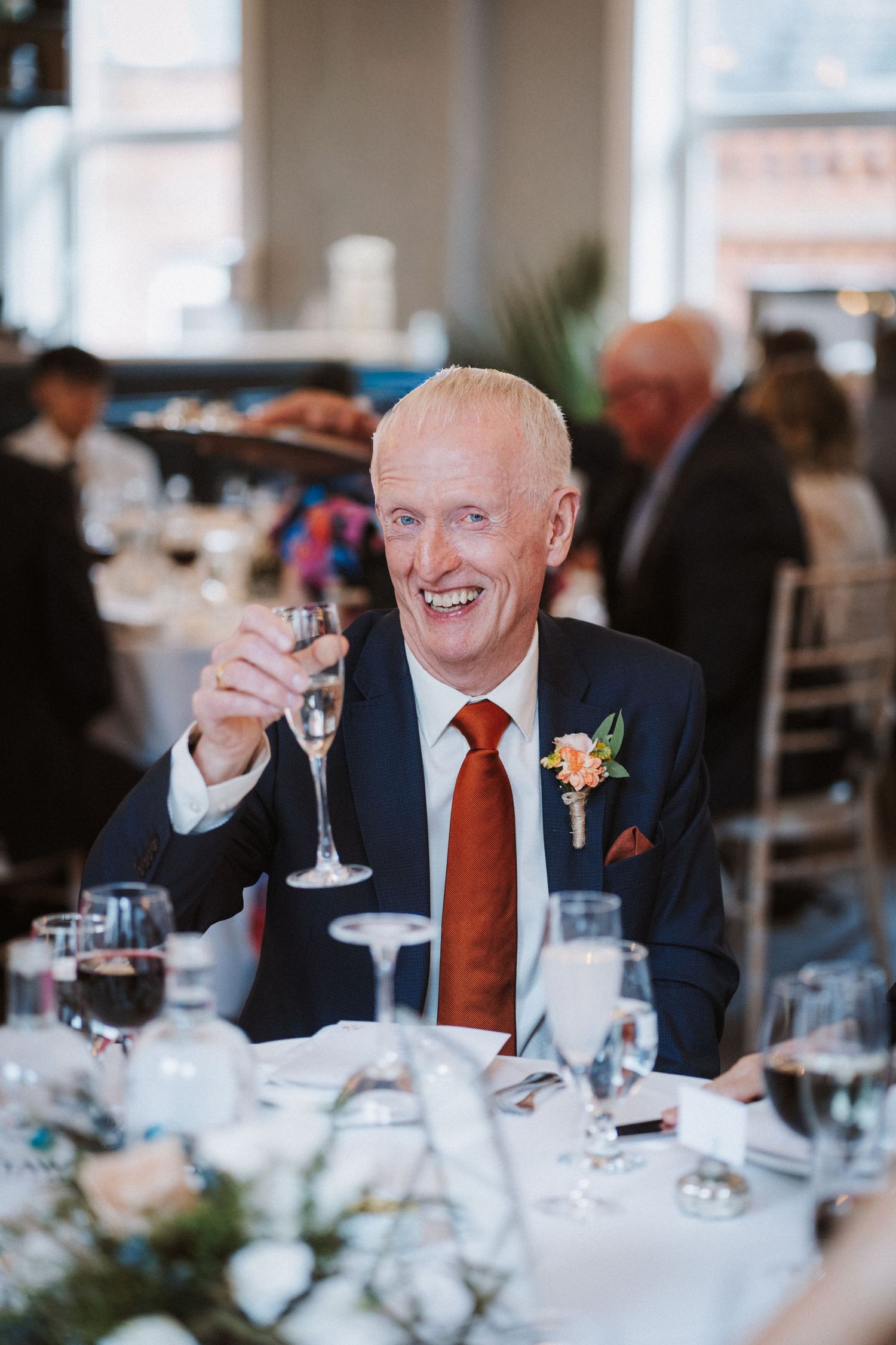 A guest in a navy suit and red tie raises a glass in celebration at an elegant wedding reception dinner.